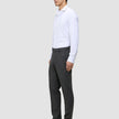 Ein Mann in einem weißen Hemd und einer grauen Motion Suit-Hose in der Farbe Regular Pepper Grey steht im Profil und trägt schwarze Slipper.