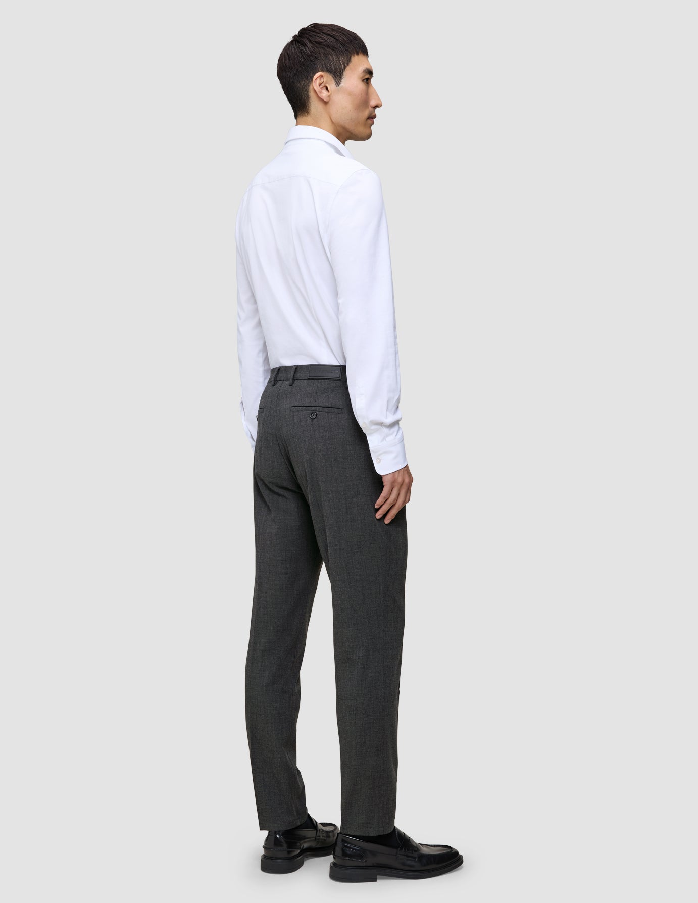Ein Mann, von hinten gesehen, der ein weißes Hemd, eine Motion Suit-Hose in der Passform Regular in der Farbe Pepper Grey und schwarze Loafer trägt.