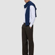 Ein Mann trägt ein weißes, fein gestreiftes Hemd mit Knöpfen, einen marineblauen Pullover, der über seinen Schultern liegt, sowie die Motion Suit Pants Relaxed Cognac – eine dunkelbraune Anzughose mit 360°-Stretch und lockerer Passform.
