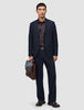Ein Mann, der eine Motion Suit Pants Relaxed in Mitternachtsblau, ein dunkles Hemd und Slipper trägt und eine braune Lederaktentasche in der Hand hält.