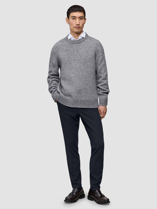 Ein Mann, der einen grauen Pullover mit Rundhalsausschnitt über einem weißen Hemd mit Kragen trägt, dazu eine Motion-Anzughose in der Farbe Slim Midnight Blue und braune Slipper.