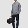 Ein Mann, der einen grauen Pullover über einem weißen Hemd mit Kragen und eine Motion Suit-Hose in der Farbe Slim Midnight Blue trägt und eine braune Reisetasche bei sich hat.