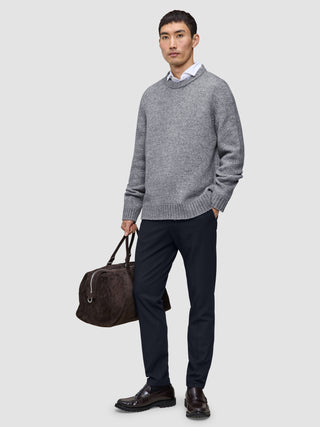 Ein Mann, der einen grauen Pullover über einem weißen Hemd mit Kragen und eine Motion Suit-Hose in der Farbe Slim Midnight Blue trägt und eine braune Reisetasche bei sich hat.