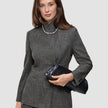 Eine Frau, die den Anzug trägt – ein grauer, karierter Nara-Blazer mit hohem Kragen und dazu passende Nara-Hose –, hält eine schwarze Clutch in der Hand.