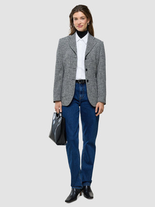 Eine Frau, die einen Tweedblazer über einem übergroßen, langärmeligen Hemd in Weiß und einem schwarzen Rollkragenpullover trägt, dazu blaue Jeans und schwarze Stiefel, und eine schwarze Handtasche bei sich hat.