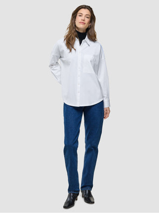 Eine Frau trägt ein weißes, übergroßes Langarmshirt über einem schwarzen Rollkragenpullover, dazu blaue Jeans und schwarze Stiefel.