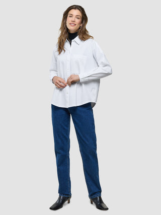 Eine Frau, die ein weißes, übergroßes Langarmshirt über einem schwarzen Rollkragenpullover trägt, dazu blaue Jeans und schwarze Stiefeletten.