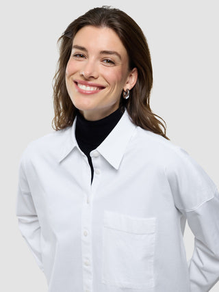 Eine lächelnde Frau, die ein weißes, übergroßes Langarmshirt über einem schwarzen Rollkragenpullover trägt.