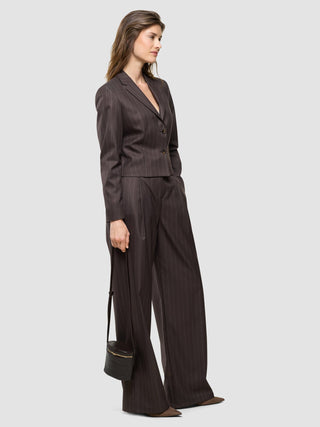 Eine Frau, die einen eleganten kurzen Damenblazer „Raina“ in Kastanienbraun mit Nadelstreifen und eine dazu passende Hose mit weitem Bein trägt und eine kleine runde Umhängetasche in der Hand hält.
