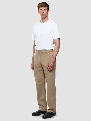 Ein Mann, der ein weißes T-Shirt und eine „Refine Cotton Pants Relaxed Fit Sand“ in Beige sowie schwarze Anzugschuhe trägt.