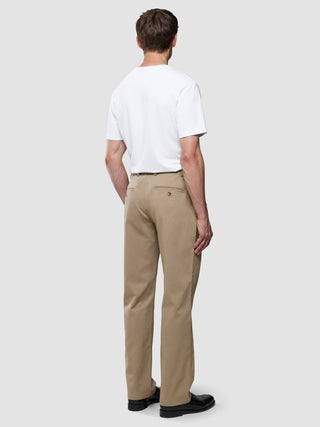 Ein Mann, von hinten gesehen, der ein weißes T-Shirt, eine „Refine Cotton Pants Relaxed Fit Sand“ und schwarze Slipper trägt.