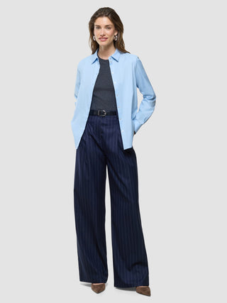 Eine Frau trägt ein hellblaues Hemd mit Knöpfen über einem dunkelgrauen melierten „Women Rib“-T-Shirt, dazu eine marineblaue, breit geschnittene Hose mit Nadelstreifen und einen schwarzen Gürtel – ein Look, der sich durch skandinavisches Design auszeichnet.