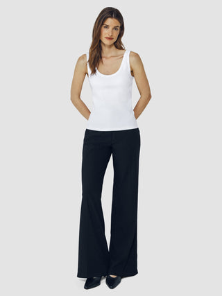 Eine Frau, die ein weißes Ripp-Tanktop und eine schwarze Hose trägt und die Hände hinter dem Rücken hat.