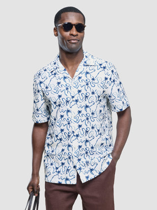 Ein Mann, der eine Sonnenbrille und ein kurzärmeliges Seersucker-Hemd in der Farbe „Navy Palms“ mit Bowlingkragen trägt.