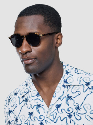 Ein Mann mit einer Sonnenbrille in Schildpattoptik und einem kurzärmeligen Seersucker-Hemd in der Farbe „Navy Palms“.