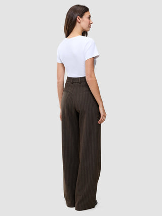 Eine Frau, von hinten gesehen, trägt ein weißes T-Shirt und eine braune Serene Pants Wide Walnut Herringbone mit weitem Bein und dezentem vertikalem Nadelstreifenmuster, die das minimalistische skandinavische Design verkörpert.