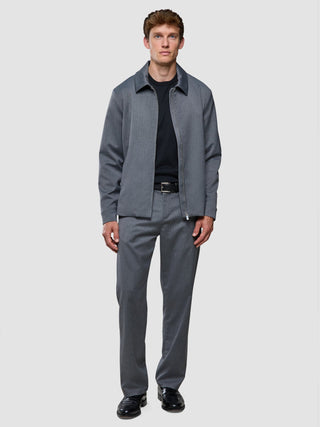 Ein Mann, der eine graue Jacke mit Kragen und Reißverschluss, ein schwarzes Hemd, die „Siena Pants Relaxed Fit Herringbone Grey“ sowie schwarze Schuhe trägt.