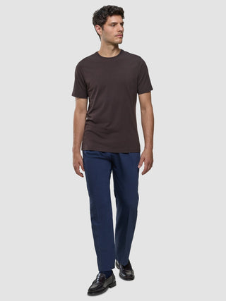 Ein Mann, der ein Supima-T-Shirt in der Farbe „Deep Brown“ und eine blaue Hose mit 360°-Stretch trägt, steht vor einem hellgrauen Hintergrund.