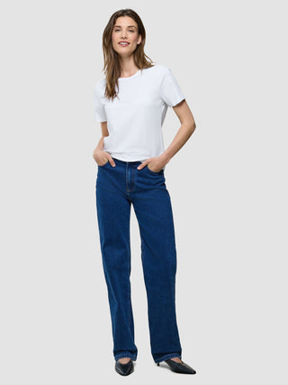 Eine lächelnde Frau, die ein weißes Supima-T-Shirt und eine blaue Jeans trägt und die Hände in den Taschen hat.