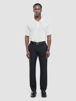 Ein Mann, der ein cremefarbenes, kurzärmeliges Strick-Polohemd, eine schwarze „Tech Linen Elastic“-Hose und schwarze Slipper trägt.