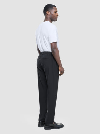 Ein Mann im Profil, der ein weißes T-Shirt, eine schwarze „Tech Linen Elastic“-Hose und schwarze Slipper trägt.
