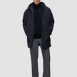 Ein Mann, der eine schwarze Utility-Jacke aus recyceltem Polyester über einem schwarzen Pullover mit Reißverschluss am Kragen und einer grauen Hose trägt.