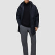 Eine Person, die eine schwarze Utility-Jacke mit Kapuze aus recyceltem Polyester über einem schwarzen Pullover mit Viertelreißverschluss, einer grauen Hose und schwarzen Anzugschuhen trägt.