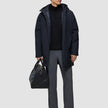 Ein Mann, der eine schwarze Utility-Jacke über einem schwarzen Pullover mit Viertelreißverschluss und einer grauen Hose trägt und eine schwarze Ledertasche in der Hand hält.