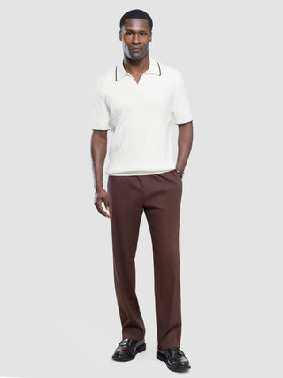 Ein Mann, der ein weißes, kurzärmeliges Polohemd mit V-Ausschnitt und eine braune Hose trägt, steht vor einem einfarbigen Hintergrund.
