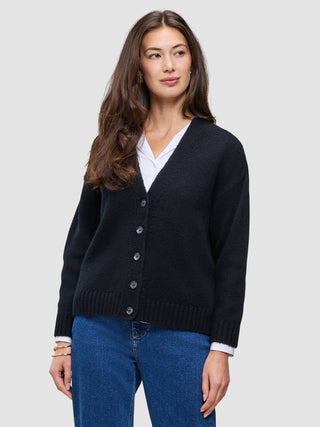 Eine Frau, die eine schwarze, grob gestrickte, weiche Wollstrickjacke für Damen über einem weißen Hemd mit Kragen und einer blauen Jeans trägt.
