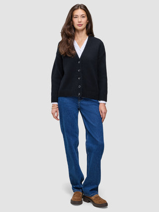 Eine Frau trägt eine schwarze, grob gestrickte, weiche Wollstrickjacke für Damen über einem weißen Hemd, dazu blaue Jeans und braune Slipper.
