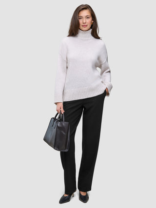 Eine Frau, die einen beigen Rollkragenpullover und eine schwarze Women GO2 Pants Straight trägt und eine schwarze Handtasche in der Hand hält.
