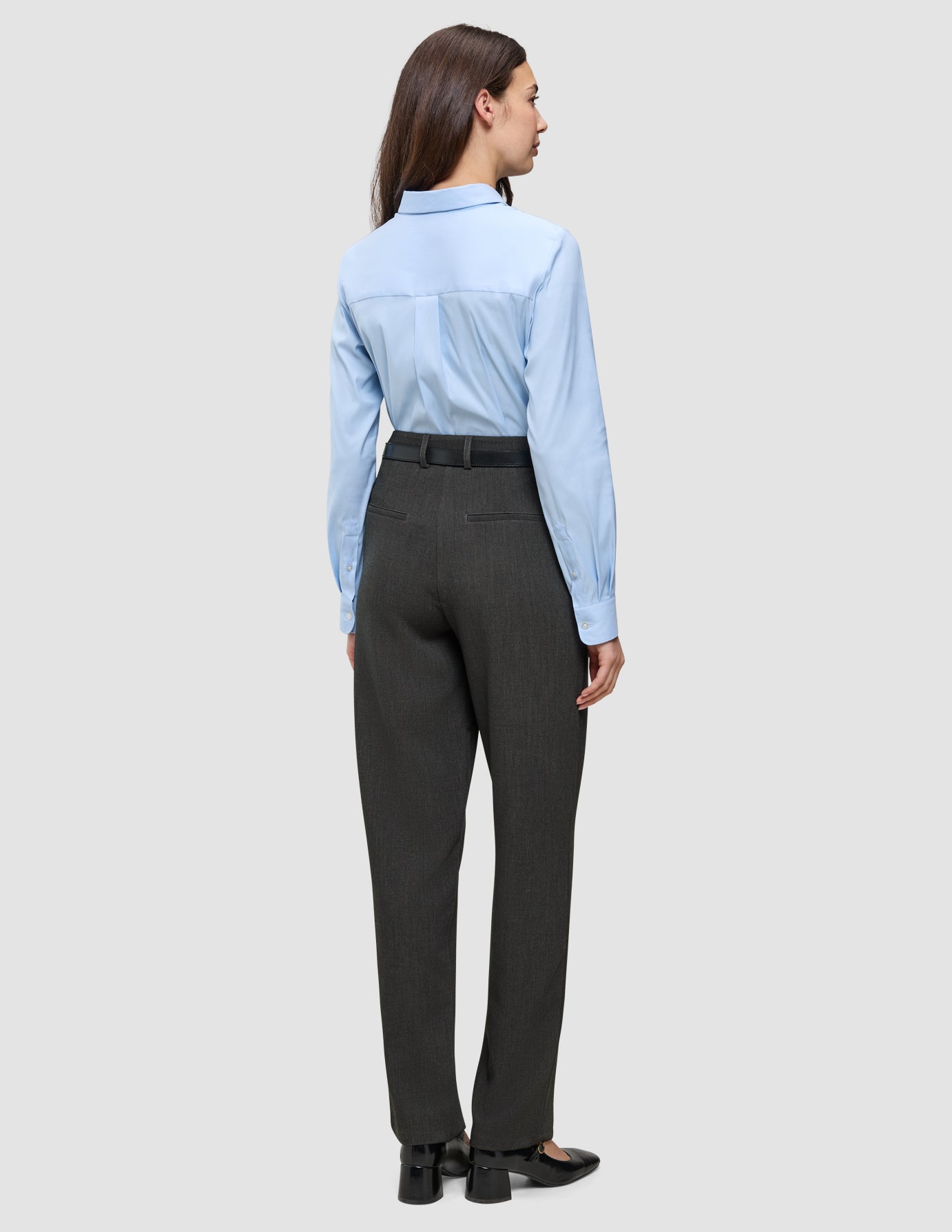 Eine Frau, von hinten gesehen, die ein hellblaues Hemd mit Knöpfen trägt, das in eine Women GO2 Pants Straight in Dunkelgrau-Melange gesteckt ist, dazu einen schwarzen Gürtel und Schuhe mit niedrigem Absatz – ein Beispiel für klares skandinavisches Design.