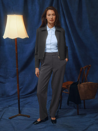 Eine Frau in einer dunkelgrauen Jacke aus Doubleface-Stoff und einem hellblauen Hemd mit Knöpfen, die eine Women GO2 Pants Straight Dark Grey Melange trägt und vor einem blauen Hintergrund steht.