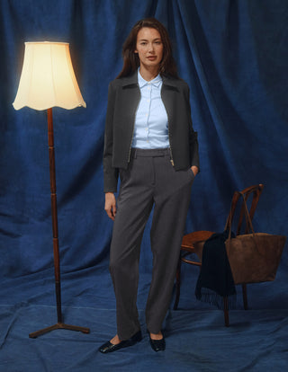 Eine Frau in einer dunkelgrauen Jacke aus Doubleface-Stoff und einem hellblauen Hemd mit Knöpfen, die eine Women GO2 Pants Straight Dark Grey Melange trägt und vor einem blauen Hintergrund steht.