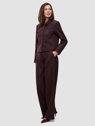 Eine Frau trägt die Women Serene Short Jacket Berry Pinstripe, eine kastenförmige, dunkelviolette Jacke mit Nadelstreifen, kombiniert mit einer passenden Hose mit hoher Taille im schlichten, skandinavischen Design.