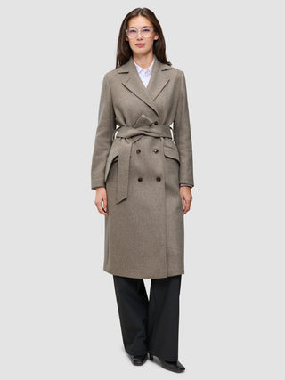 Eine Person, die einen graubeigen Damenmantel aus Wolle trägt – ein zweireihiges, mit einem Gürtel versehenes, minimalistisches skandinavisches Design –, dazu ein weißes Hemd mit Kragen und eine schwarze Hose.