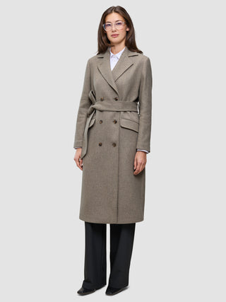 Eine Frau trägt einen Women Wool Coat Greige – einen zweireihigen Wollmantel mit Gürtel – über einem weißen Hemd und einer schwarzen Hose.