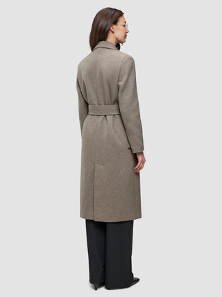 Eine Person, von hinten gesehen, die einen Women Wool Coat Greige trägt – einen langen, mit einem Gürtel versehenen, greigefarbenen Wollmantel im skandinavischen Design – über einer schwarzen Hose.