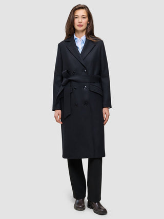 Eine Frau trägt einen schwarzen Damen-Wollmantel – einen langen, zweireihigen Wollmantel mit Gürtel in der Taille – über einem hellblauen Hemd mit Kragen und einer schwarzen Hose im klassischen skandinavischen Design.