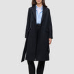 Eine Frau, die einen schwarzen Damenmantel aus Wolle über einem hellblauen Hemd mit Knöpfen und einer schwarzen Hose trägt, was das minimalistische skandinavische Design widerspiegelt.