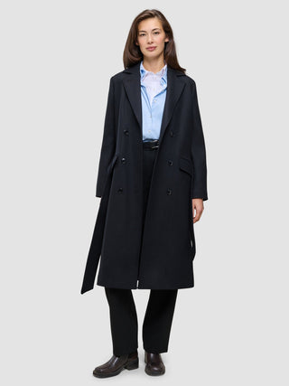 Eine Frau, die einen schwarzen Damenmantel aus Wolle über einem hellblauen Hemd mit Knöpfen und einer schwarzen Hose trägt, was das minimalistische skandinavische Design widerspiegelt.