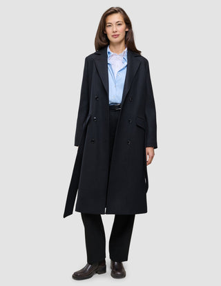Eine Frau, die einen schwarzen Damenmantel aus Wolle über einem hellblauen Hemd mit Knöpfen und einer schwarzen Hose trägt, was das minimalistische skandinavische Design widerspiegelt.