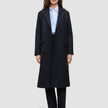 Eine Frau trägt einen schwarzen Damen-Wollmantel in knielanger Länge im Zweireiher-Stil über einem hellblauen Hemd mit Knöpfen und einer schwarzen Hose, was das klare skandinavische Design unterstreicht.