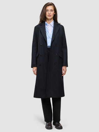 Eine Frau trägt einen schwarzen Damen-Wollmantel in knielanger Länge im Zweireiher-Stil über einem hellblauen Hemd mit Knöpfen und einer schwarzen Hose, was das klare skandinavische Design unterstreicht.
