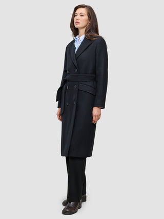 Eine Person, die einen schwarzen Damenmantel aus Wolle mit Gürtel an der Taille trägt, dazu ein hellblaues Hemd mit Kragen und eine schwarze Hose im schlichten, skandinavischen Design.
