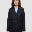Eine Frau trägt einen schwarzen Damen-Wollmantel im Zweireiher-Stil mit Bindegürtel über einem hellblauen Hemd mit Kragen im skandinavischen Design.