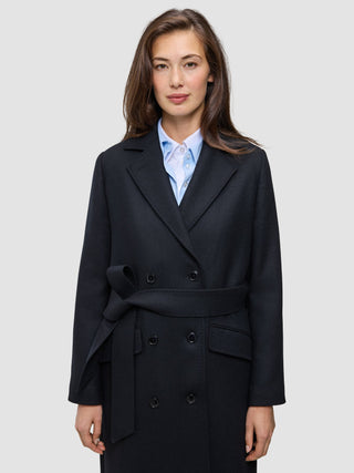 Eine Frau trägt einen schwarzen Damen-Wollmantel im Zweireiher-Stil mit Bindegürtel über einem hellblauen Hemd mit Kragen im skandinavischen Design.
