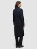 Eine Frau wird von hinten gezeigt. Sie trägt einen schwarzen Damenmantel aus Wolle in minimalistischem skandinavischem Design mit einer langen Silhouette und Gürtel.