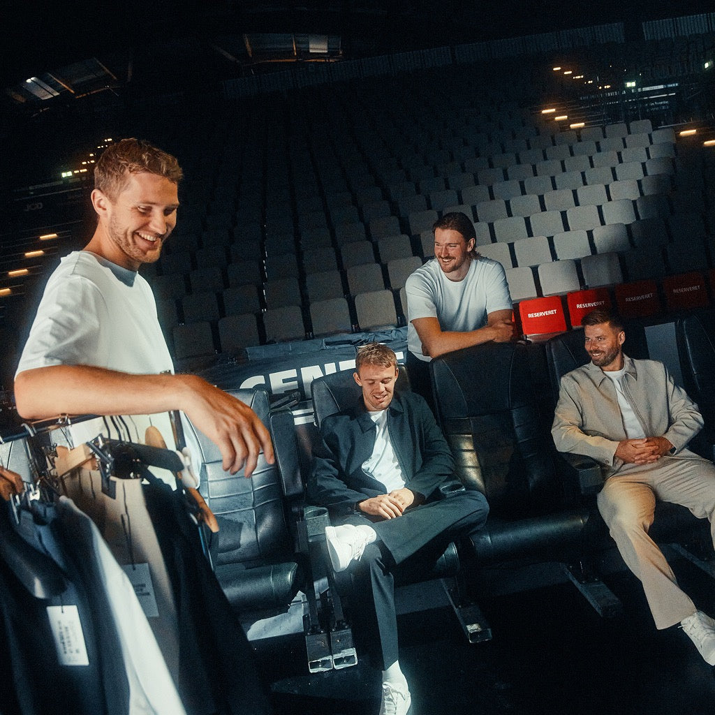 Vier Männer in einem leeren Stadiontheater; ein Mann steht im Vordergrund und hält einen Kleiderständer in der Hand, während die anderen drei im Zuschauerbereich sitzen und stehen.